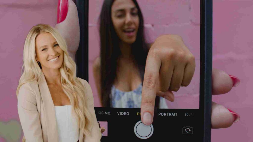 Smiling blonde woman in a beige blazer beside a giant smartphone screen showing a finger about to press the shutter button.
