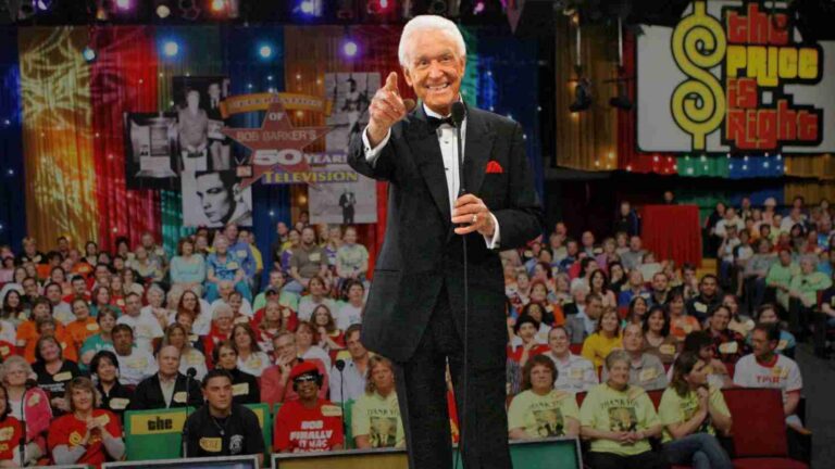Male host in a tuxedo on stage, pointing at the camera with a microphone, as a large colorful audience watches behind him on The Price Is Right set.