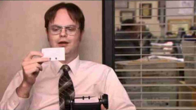 Office worker with glasses holds up a small white card toward the camera, seated at a desk behind window blinds.