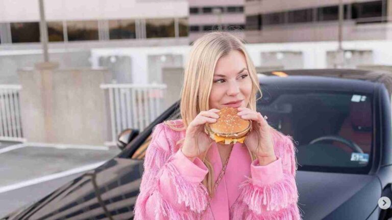 Woman in a pink fringed jacket eating a hamburger outdoors beside a parked car, smiling slightly.