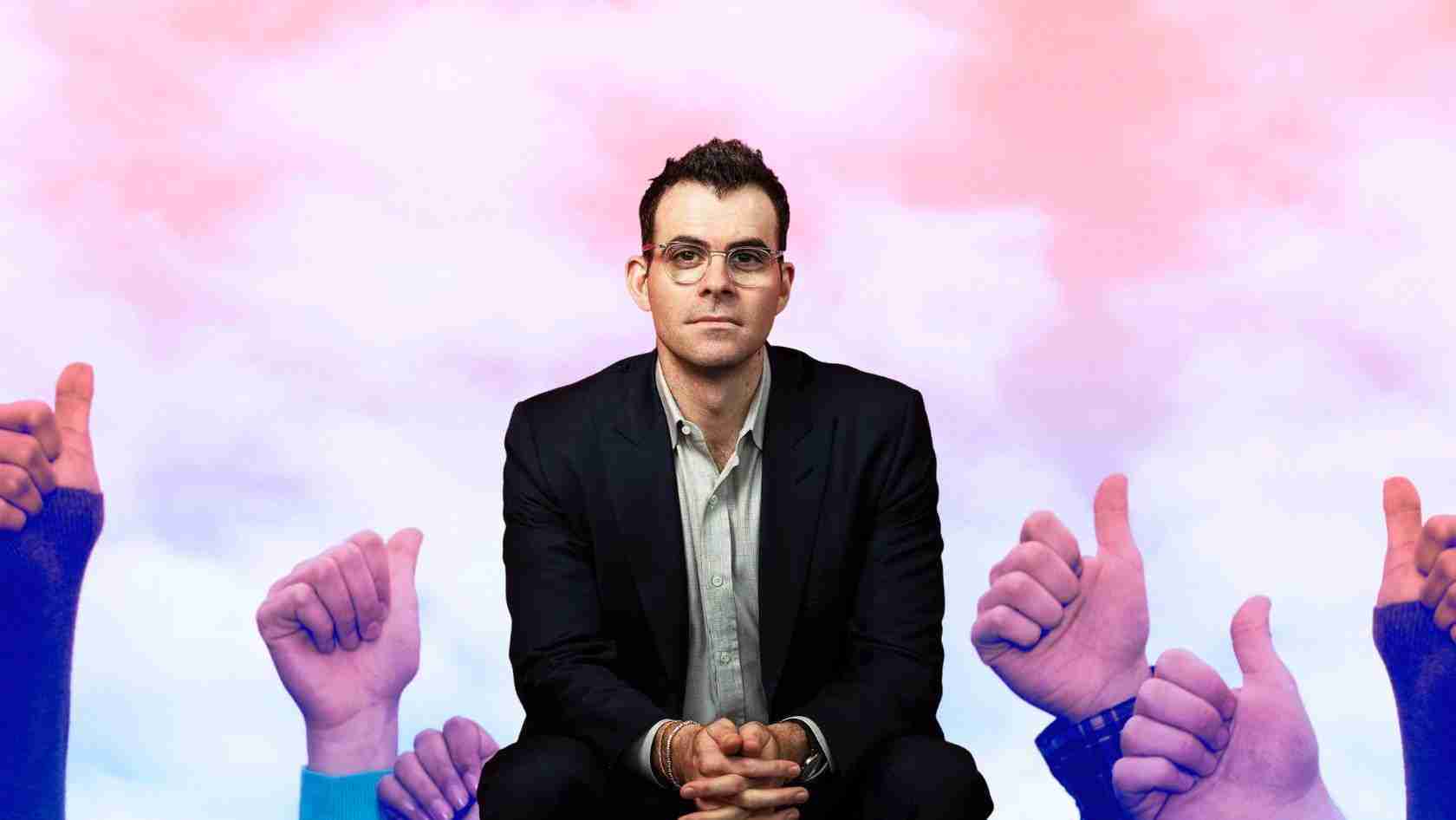 Centered man in a dark suit sits with hands clasped, surrounded by numerous thumbs-up signs against a pink-to-blue gradient background, signaling approval or endorsement.