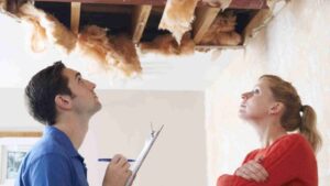 Man with a clipboard explains renovation to a woman as they inspect a ceiling with exposed insulation and torn drywall/sawdust above them.