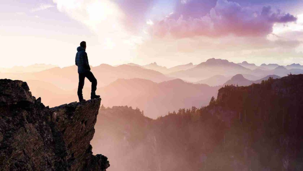 Person standing on a rocky cliff edge overlooking layered mountains at sunrise, panoramic view in soft pastel colors