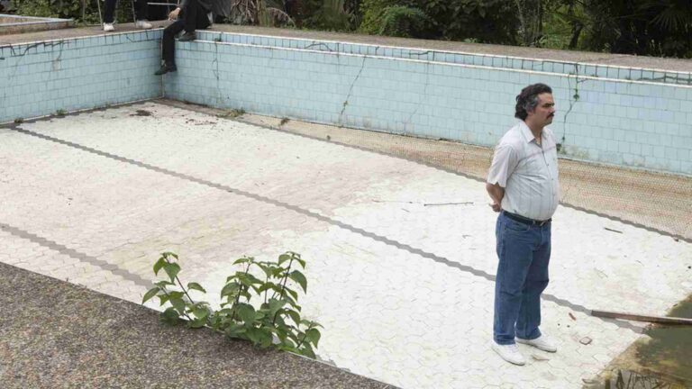 Man with mustache in a light shirt and jeans stands with hands behind his back beside an empty, cracked swimming pool.
