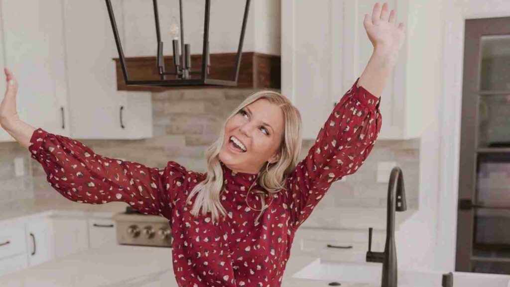 Joyful woman in a red patterned blouse stretches arms up with a big smile in a bright kitchen.