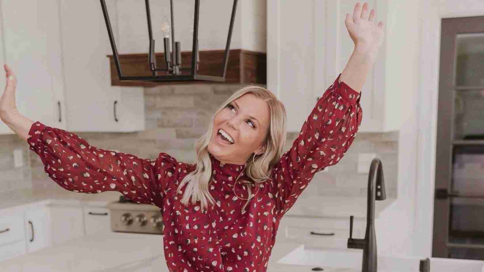 Joyful woman in a red patterned blouse stretches arms up with a big smile in a bright kitchen.