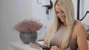 Smiling woman with long blonde hair sits on a couch, using a smartphone in a light, cozy living room.