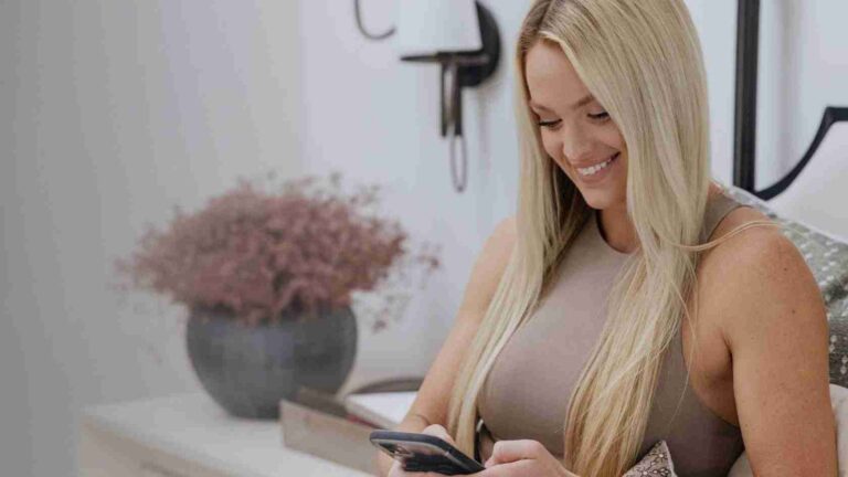 Smiling woman with long blonde hair sits on a couch, using a smartphone in a light, cozy living room.