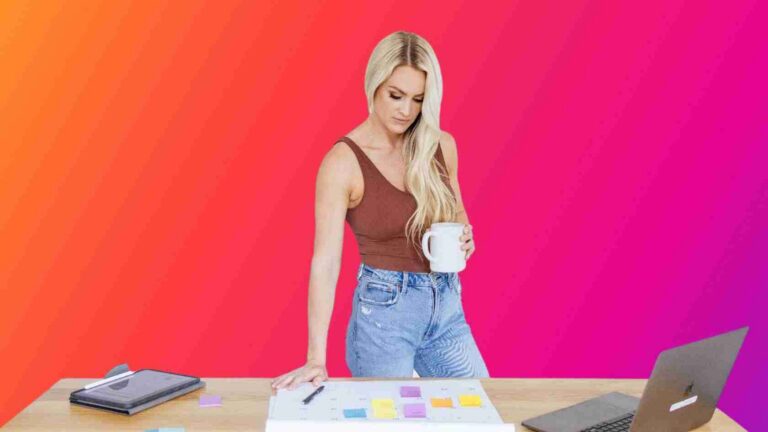 Woman standing at a desk, organizing colorful sticky notes and papers while holding a white mug, with a laptop nearby.