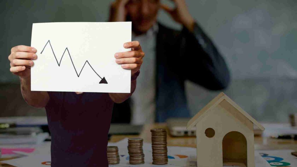 Person holding a white chart with a downward line, coins stacked nearby, and a small house model on a desk.