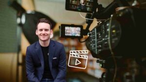 Smiling man in a navy blazer sits in a filming studio with a professional video camera in the foreground.