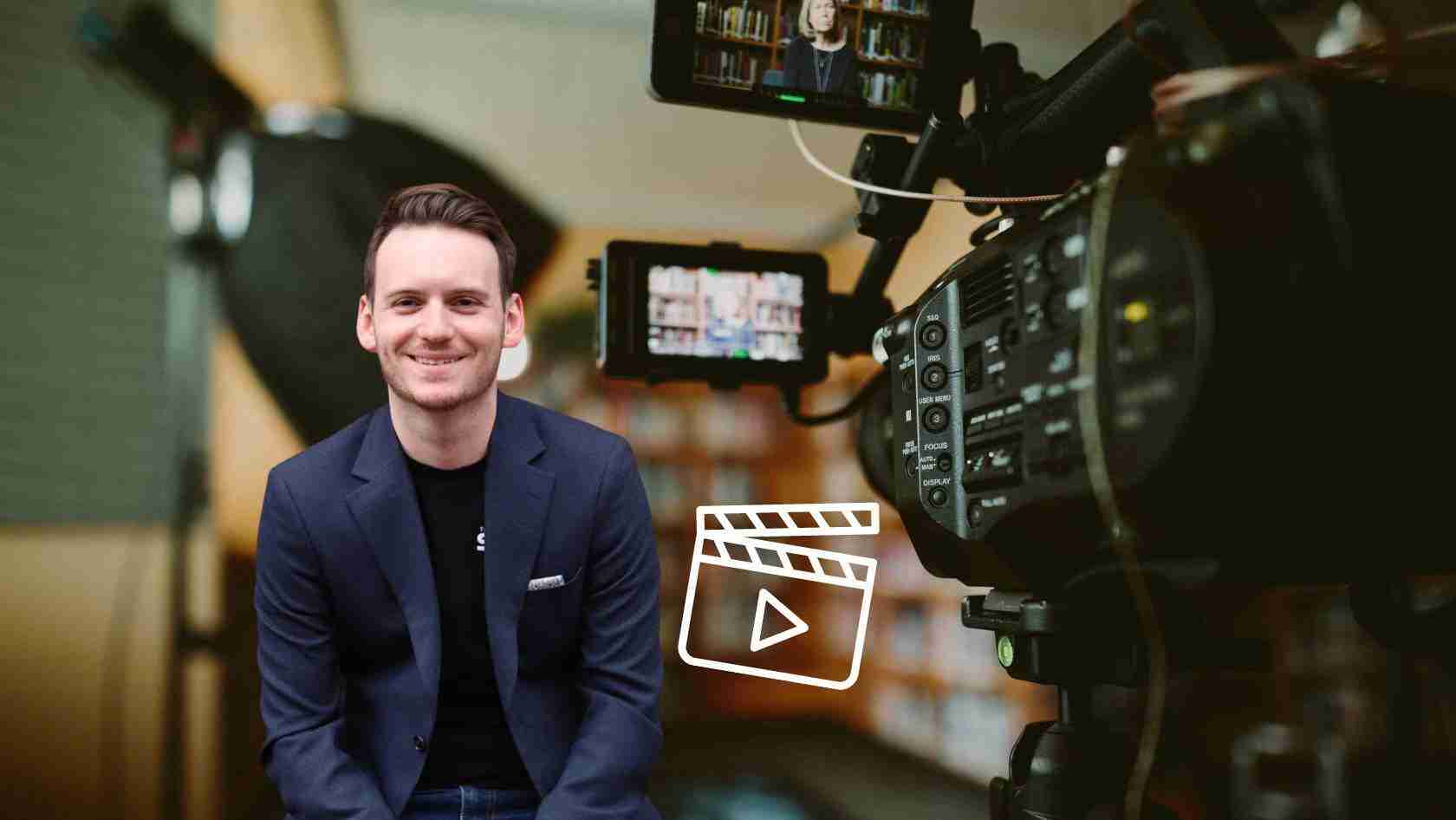 Smiling man in a navy blazer sits in a filming studio with a professional video camera in the foreground.