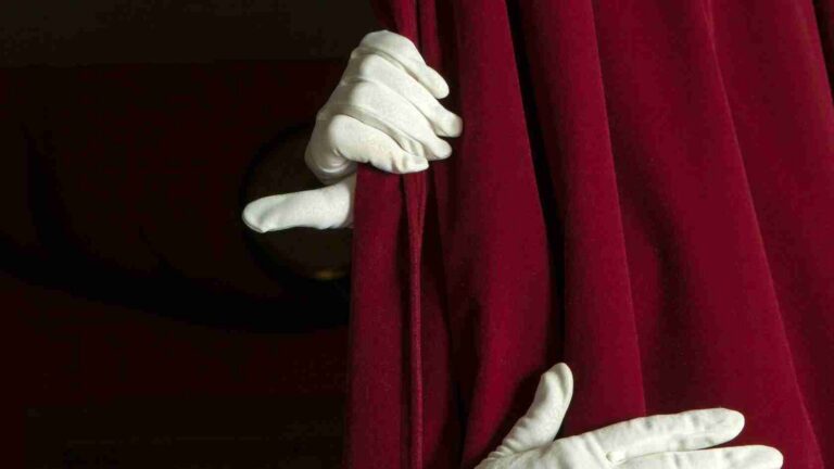 White-gloved hands pull a deep red velvet theater curtain aside, revealing darkness behind it.