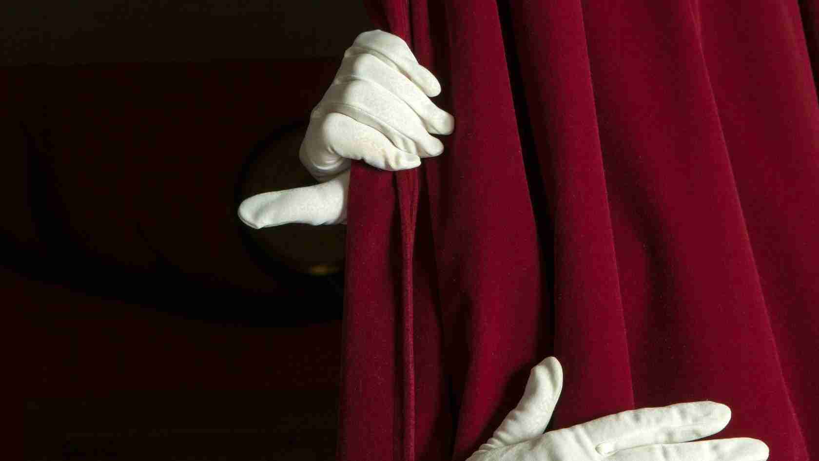 White-gloved hands pull a deep red velvet theater curtain aside, revealing darkness behind it.