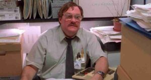 Middle-aged man in a green shirt and tie sits at a cluttered office desk with papers and folders around him, wearing glasses and a name badge.