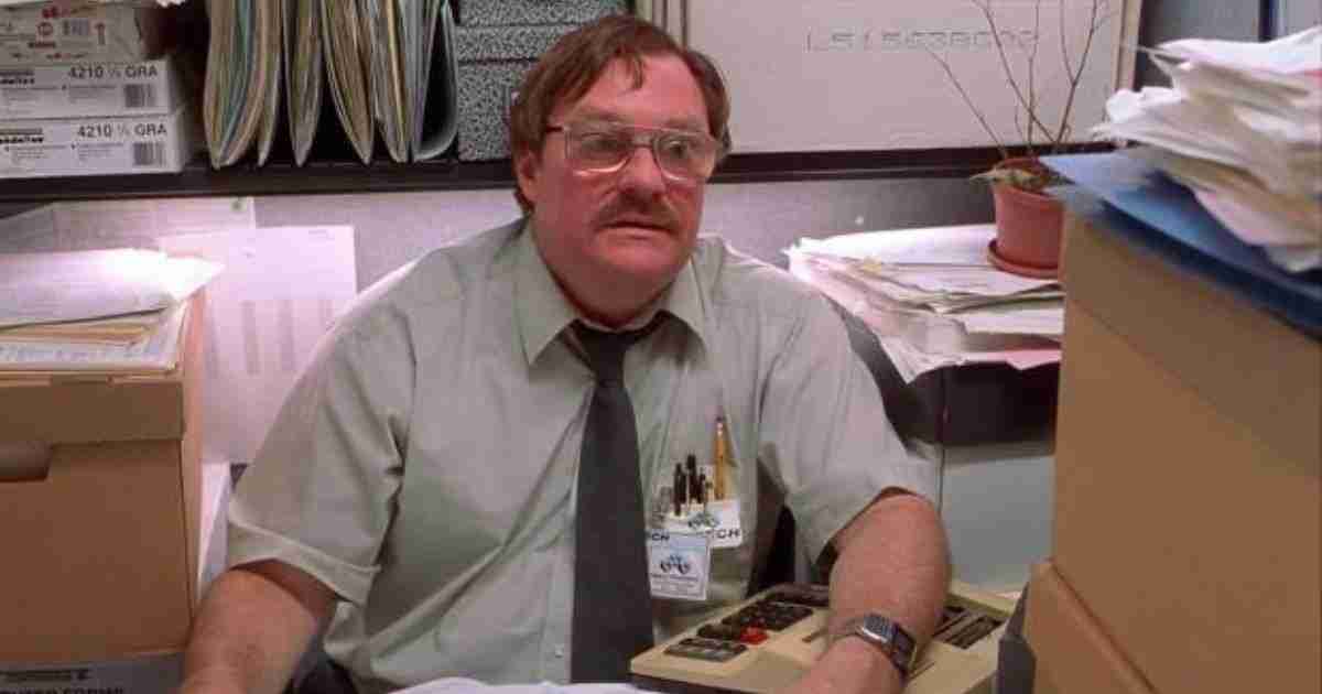 Middle-aged man in a green shirt and tie sits at a cluttered office desk with papers and folders around him, wearing glasses and a name badge.