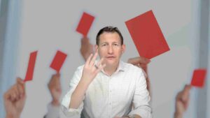 Man in a white shirt seated at a table, surrounded by hands holding red cards.