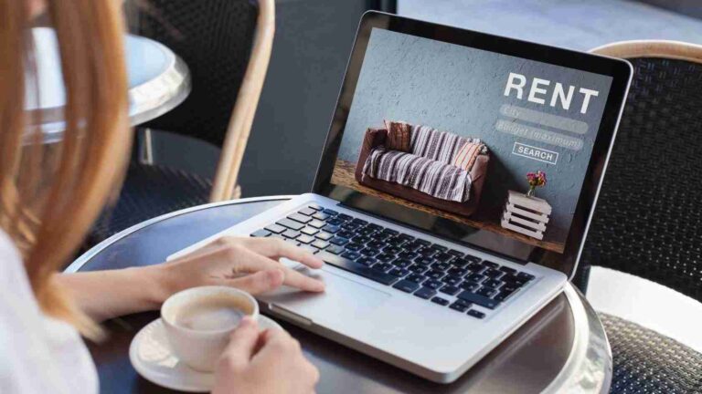 Person using a laptop at an outdoor table, the screen shows a furniture rental page with a sofa and the word RENT and a SEARCH button.