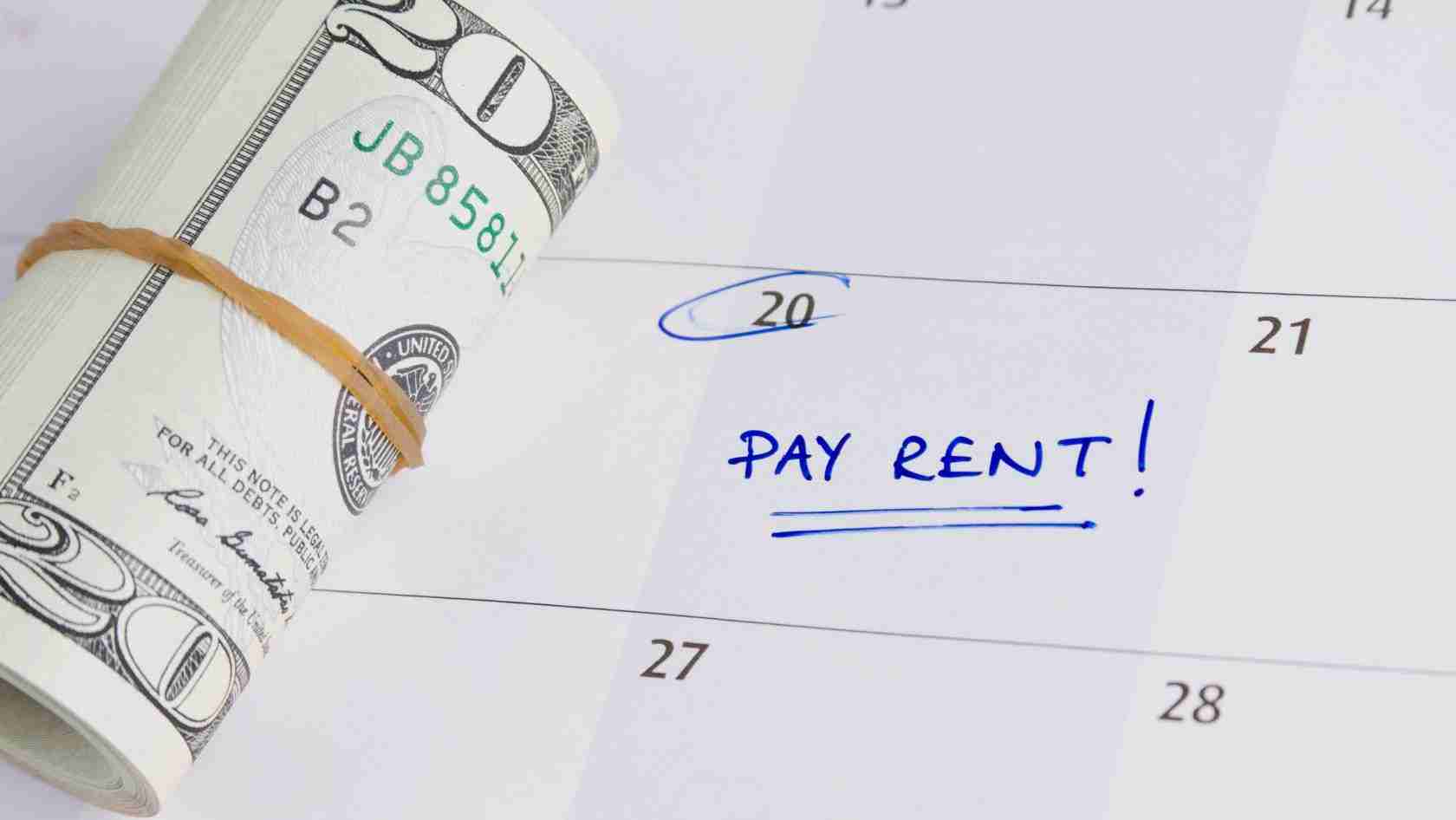 Rolled bundle of U.S. dollar bills with a rubber band on a calendar showing 'PAY RENT!' written in blue ink.