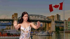 Woman in a floral dress raises both hands in front of a marina, with boats, a bridge, and a waving Canadian flag nearby.