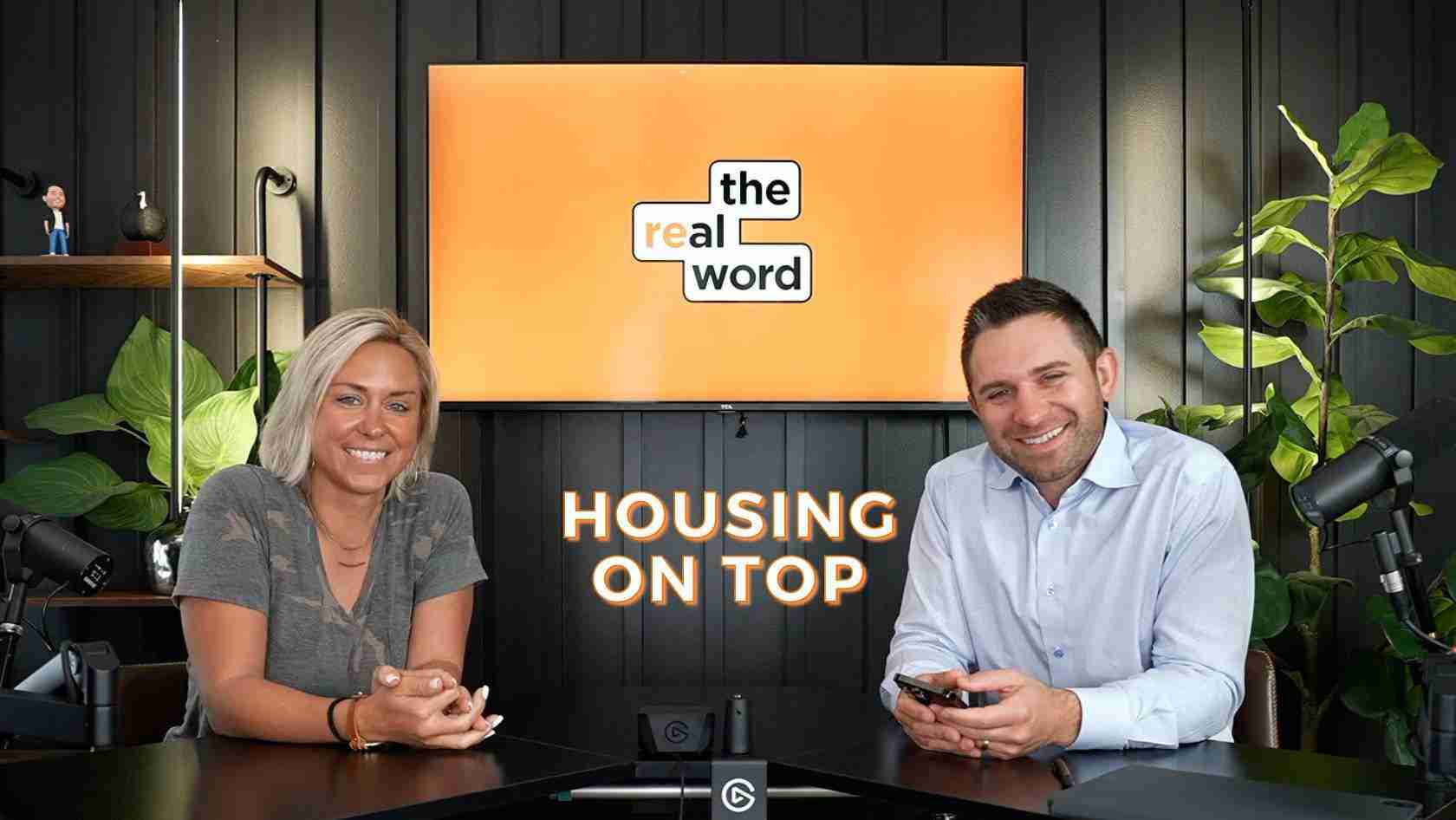 Two hosts sit at a dark studio desk in front of a bright orange screen reading 'the real word' and 'HOUSING ON TOP'.