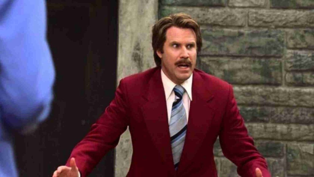 Man in a burgundy suit and striped tie, arms outstretched, shouting at someone off‑camera against a stone wall.