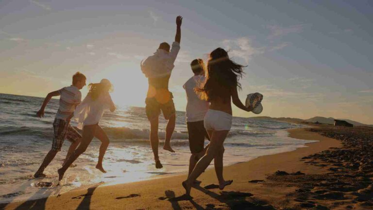 Group of friends jumping along a sunny beach at sunset, with waves and footprints in the sand.