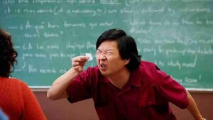 Man in a red shirt scrunches his face and holds a tissue up to his eye in a classroom with a chalkboard full of equations behind him.