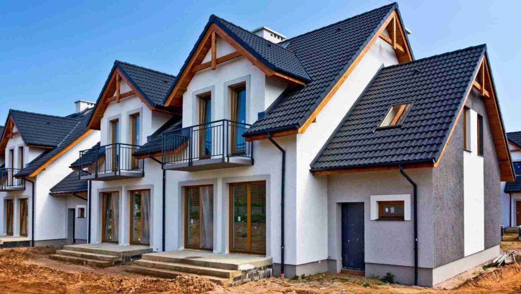 Row of white modern townhouses with dark blue tiled roofs, wooden eaves, and glass patio doors under construction.