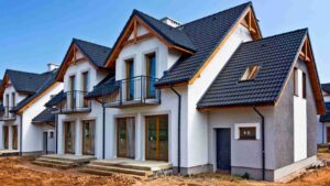 Row of white modern townhouses with dark blue tiled roofs, wooden eaves, and glass patio doors under construction.