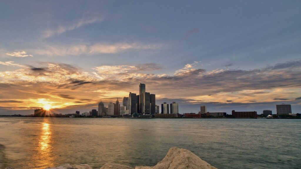 Sunset over a city skyline across a calm body of water, with rocks in the foreground.