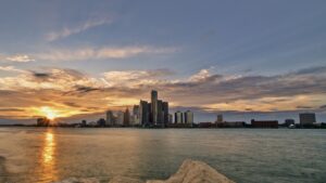 Sunset over a city skyline across a calm body of water, with rocks in the foreground.