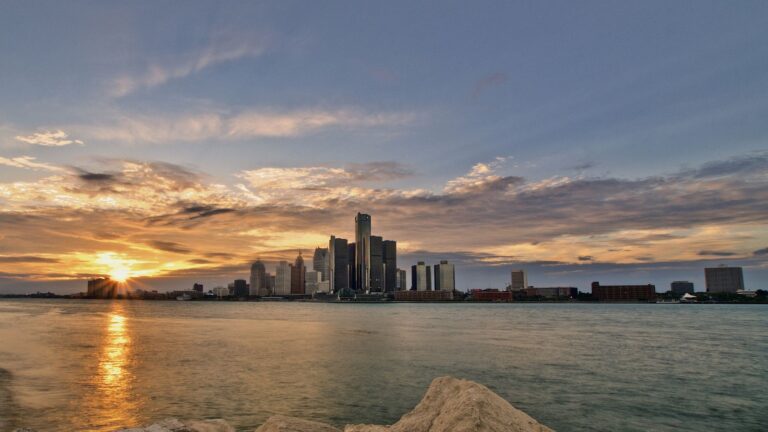 Sunset over a city skyline across a calm body of water, with rocks in the foreground.