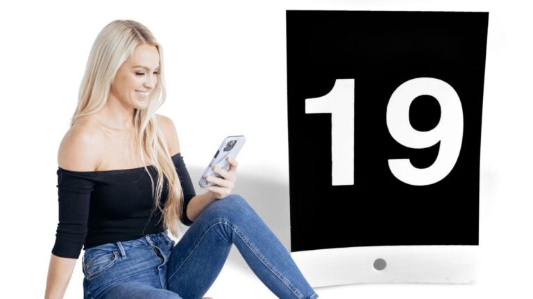 Woman sitting in jeans and a black off-the-shoulder top, looking at her phone, beside a large black sign with the white number 19
