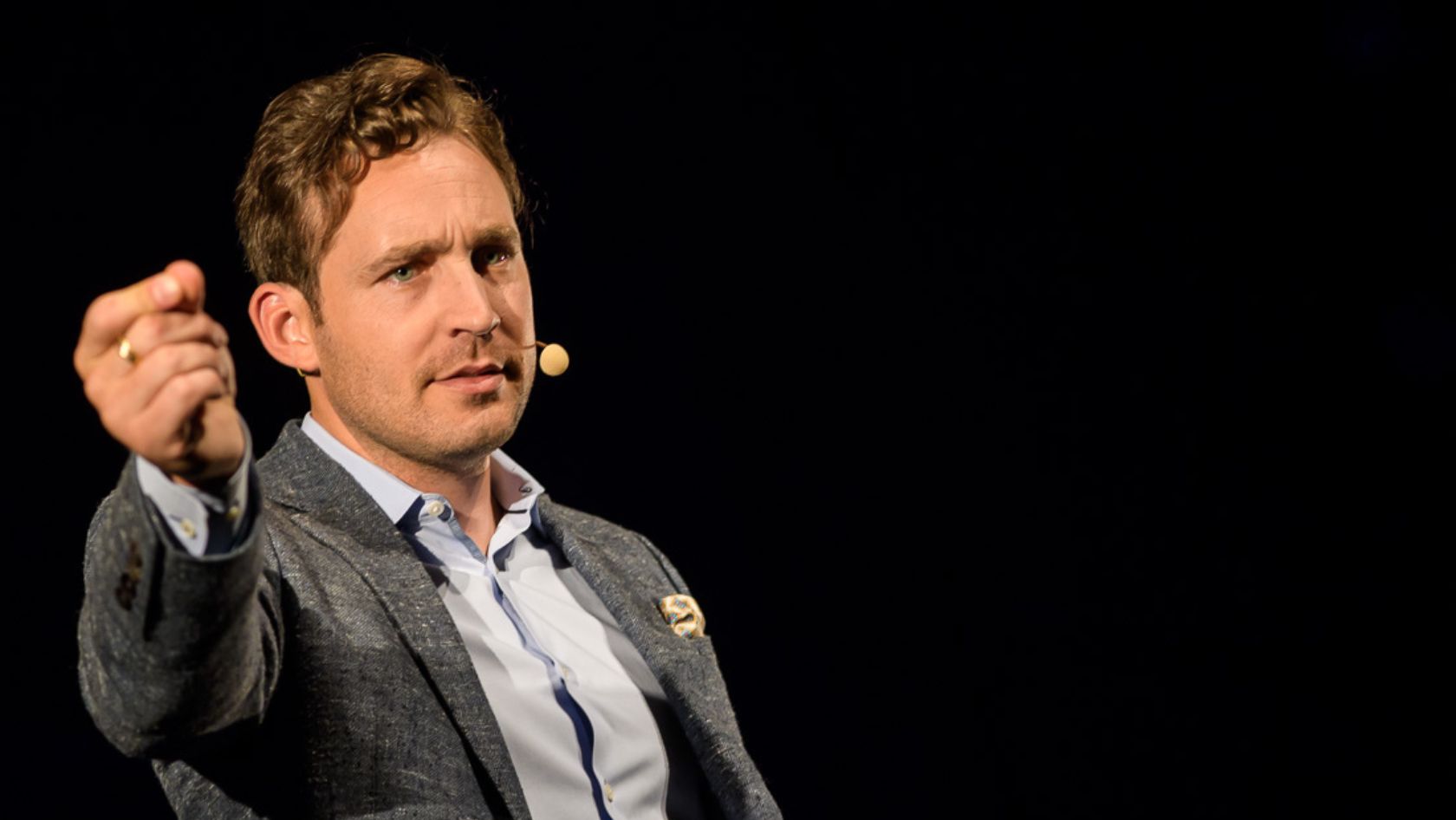 Male presenter on a dark stage, wearing a gray blazer and light shirt, pointing toward the audience with a headset microphone visible.