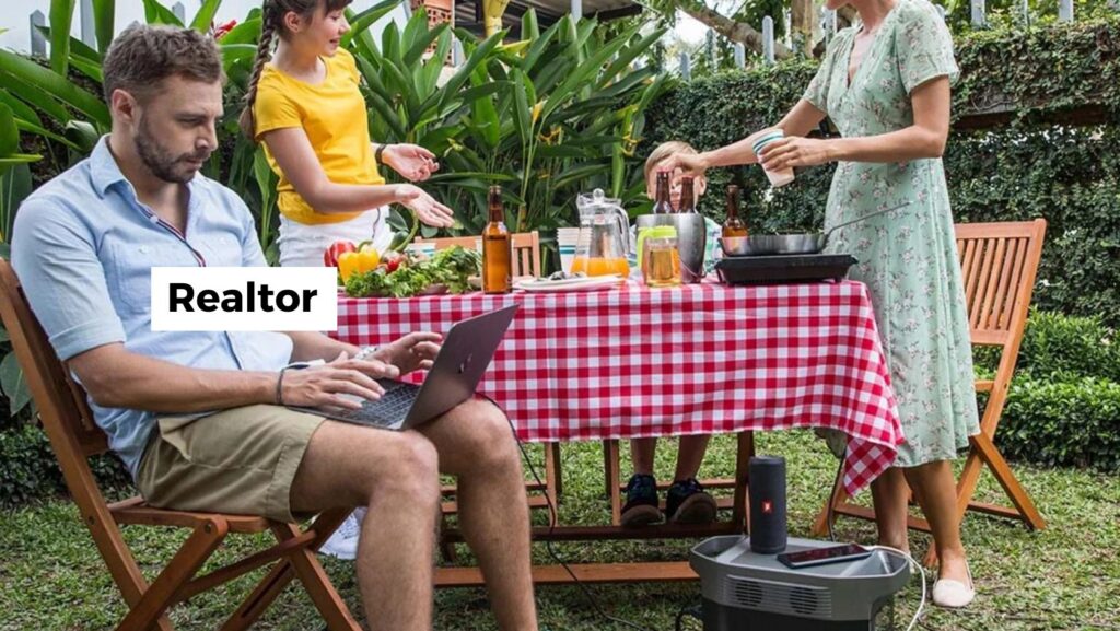 Man with a laptop seated at a red‑and‑white checkered tablecloth in a sunny backyard while two women grill and prep food nearby.