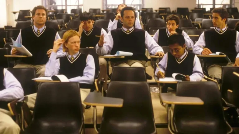 Students in matching vests and ties sit in a college lecture hall with individual desks; a center student gestures as if asking a question.