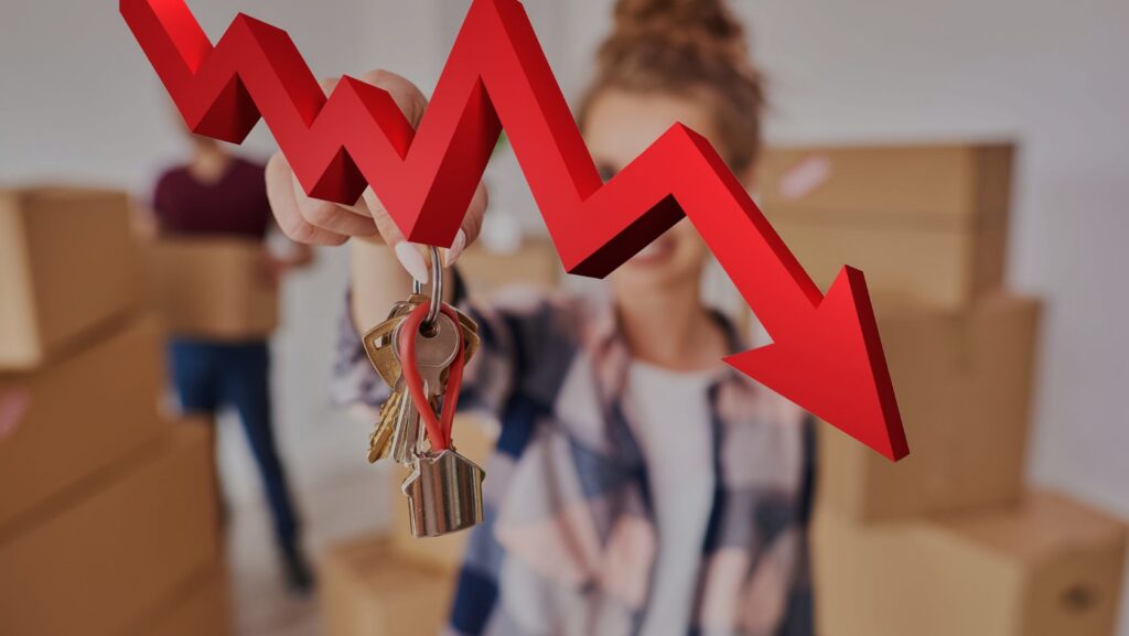 Person holds a set of keys toward the camera with a large red downward-arrow graph in the foreground, suggesting falling housing prices or a downturn.