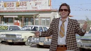 Man in a brown plaid suit and sunglasses gesturing in front of a used car lot with vintage cars selling for cheap.
