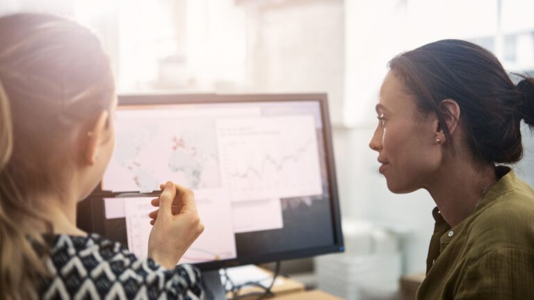 Two colleagues in an office reviewing charts on a computer monitor, one holding a pen and pointing at the screen.