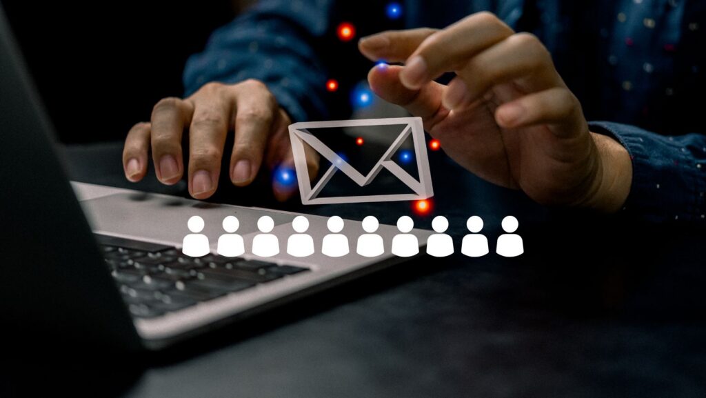 Hands over a laptop with a paper envelope icon between them, and a row of user icons below, symbolizing email or phishing targeting many people.