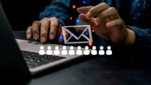 Hands over a laptop with a paper envelope icon between them, and a row of user icons below, symbolizing email or phishing targeting many people.