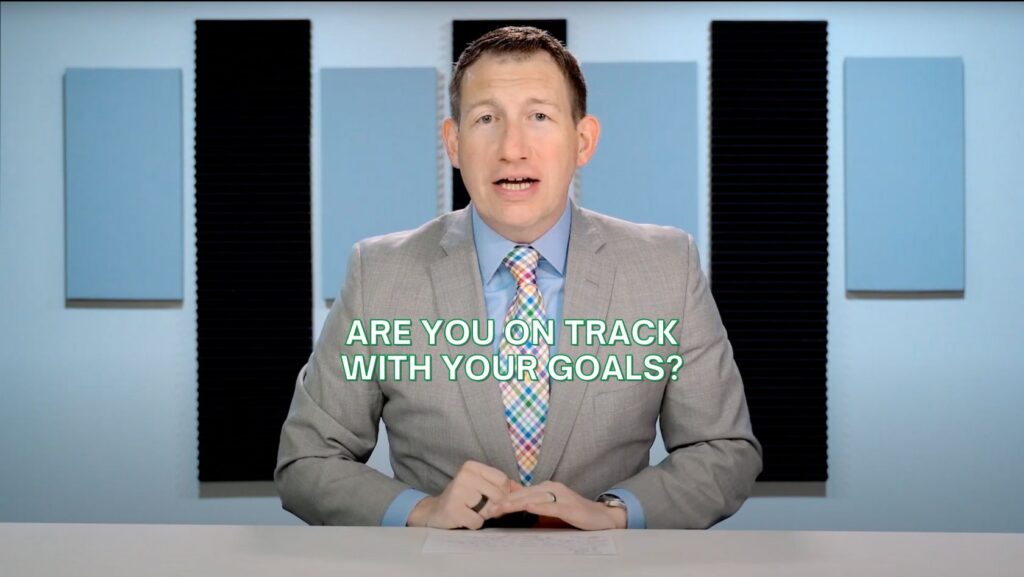 Professional presenter in a gray suit at a desk, studio background, with the caption 'ARE YOU ON TRACK WITH YOUR GOALS?'