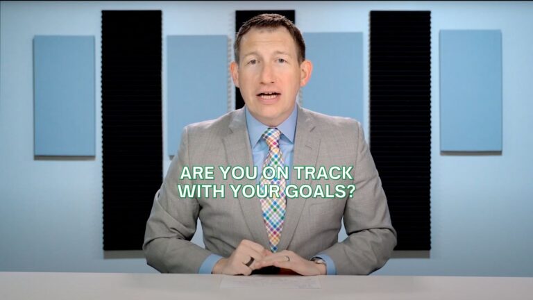 Professional presenter in a gray suit at a desk, studio background, with the caption 'ARE YOU ON TRACK WITH YOUR GOALS?'