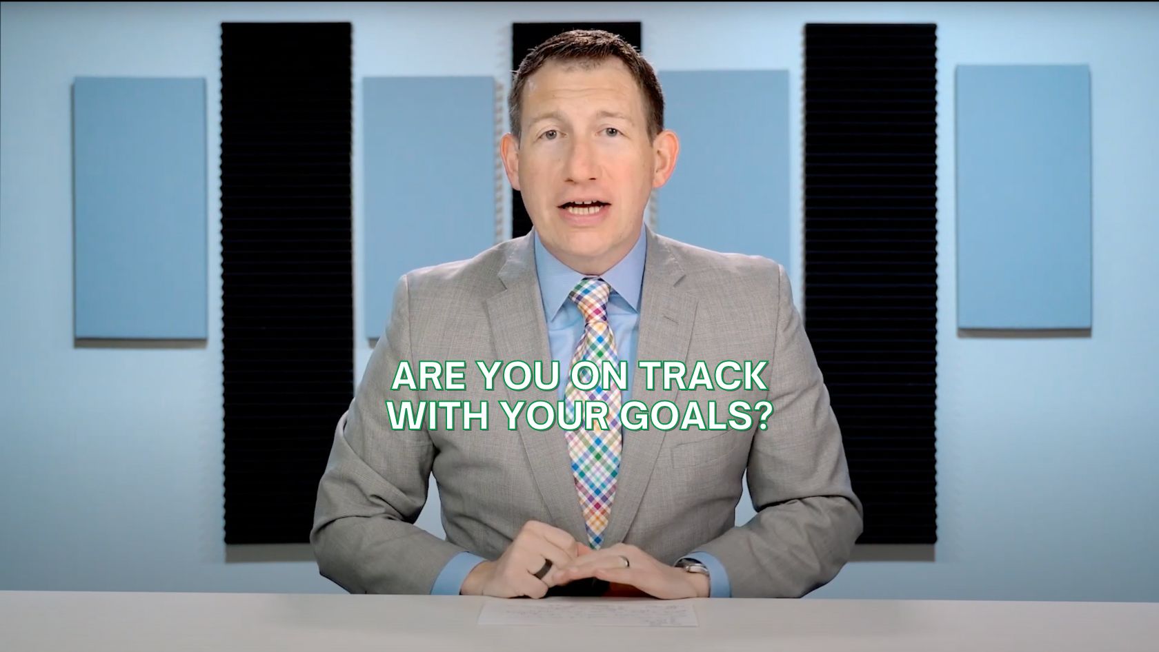 Professional presenter in a gray suit at a desk, studio background, with the caption 'ARE YOU ON TRACK WITH YOUR GOALS?'