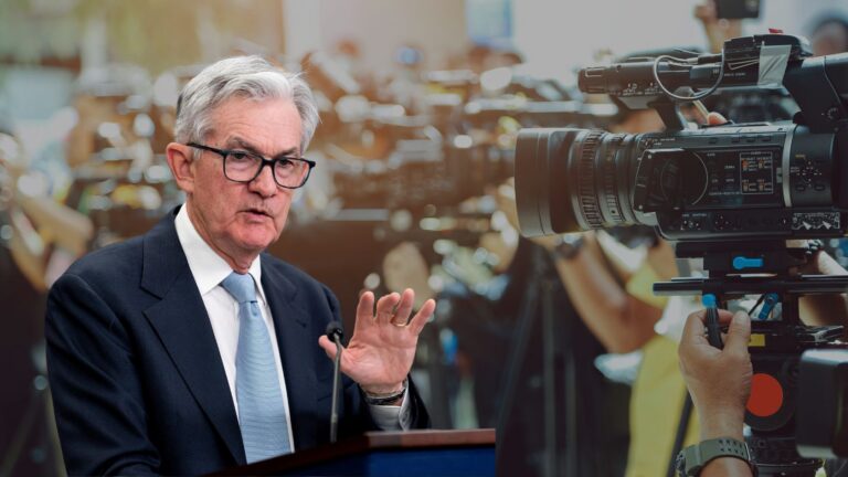 Man in a suit and glasses speaks at a podium during a press conference as cameras film in the background.