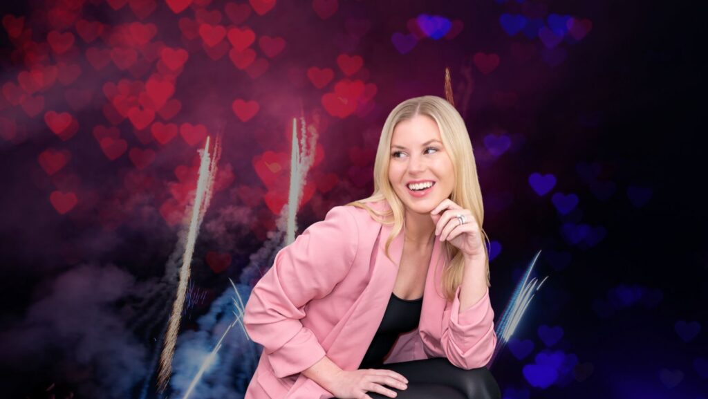 Smiling blonde woman in a pink blazer seated against a background of pink heart-shaped bokeh lights and fireworks.