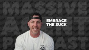 Smiling man in a white T‑shirt and backward black cap, with bold 'Embrace the Suck' text on a dark patterned background.