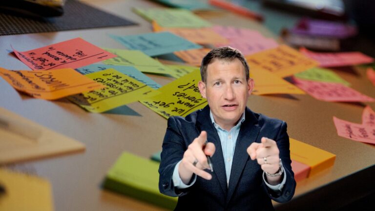 Man in a suit points at the camera with colorful sticky notes scattered on a conference table behind him.