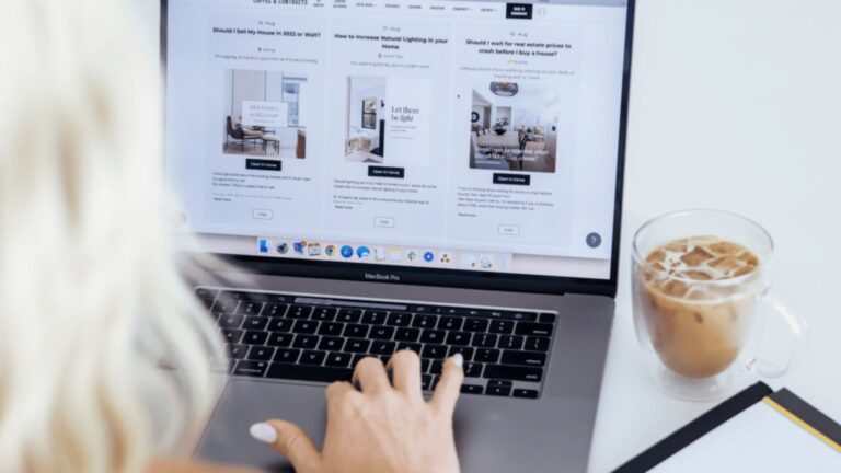Person typing on a MacBook Pro with a three-column website layout on screen; iced coffee beside.